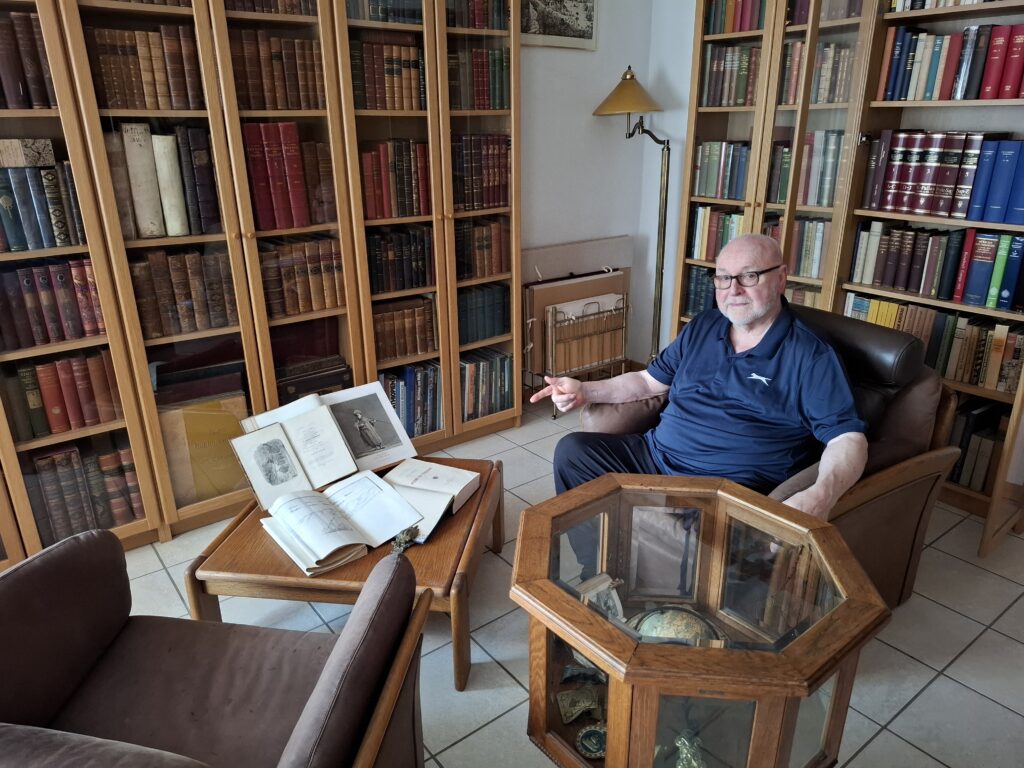 Mike sitting in his private library pointing to open books on a coffee table. Mike, M. H. Broderick, sitting in his private library pointing to open books on a coffee table.